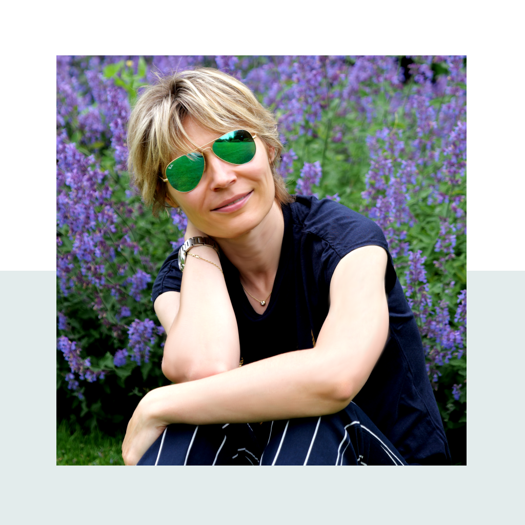 Anna sitting in a field of purple flowers, wearing green aviator sunglasses and smiling