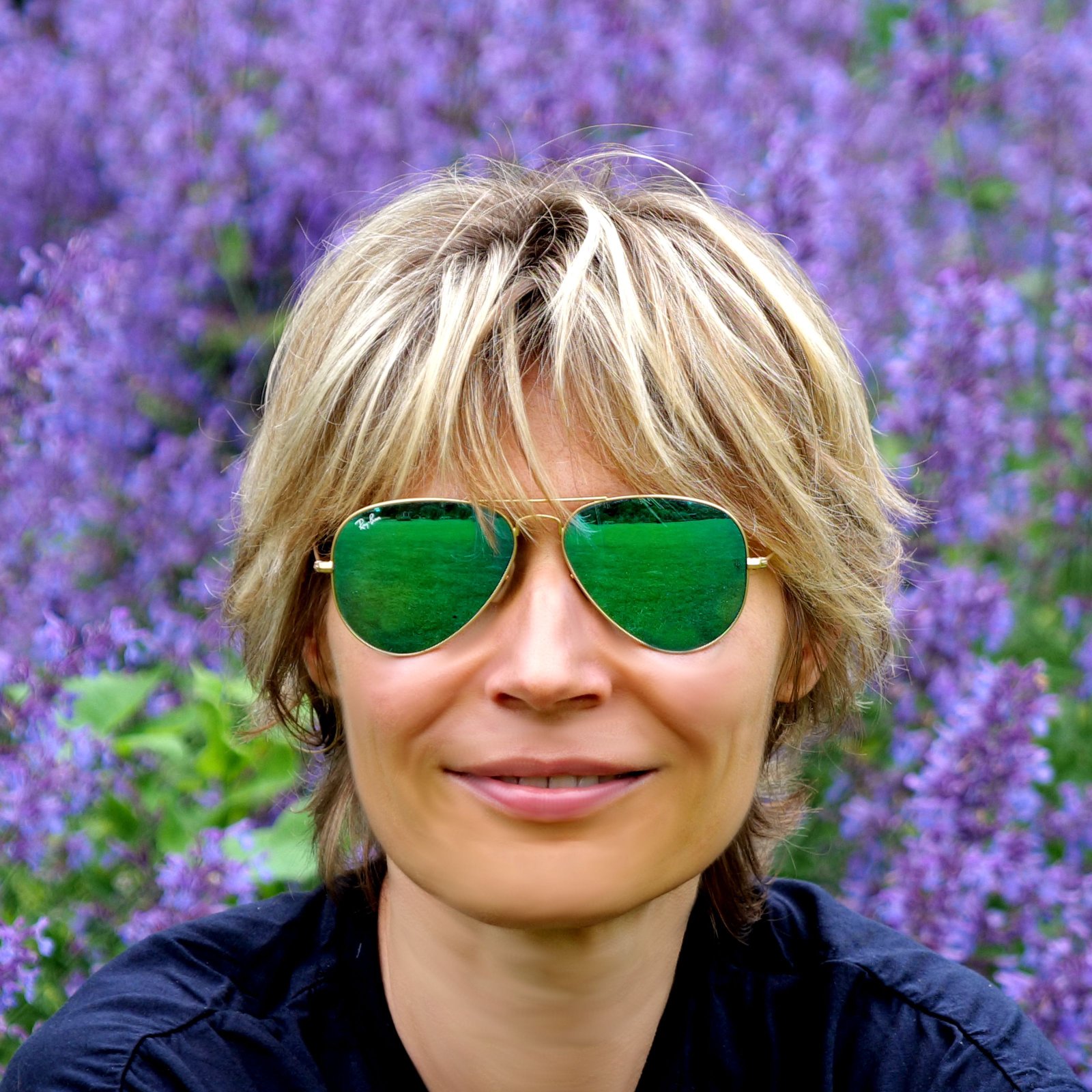 Portrait of Anna wearing green aviator sunglasses in front of purple flowers