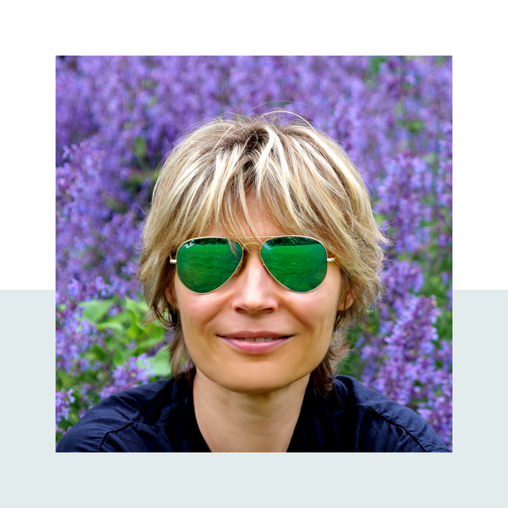 Anna smiling, wearing green aviator sunglasses, in front of a field of purple flowers